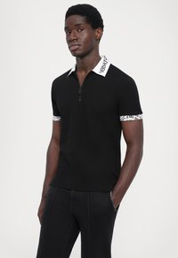 Svart polo-skjorte med kort glidelås, hvit krage med logo-tekst og mønstrede detaljer på ermene. Har en tett passform.