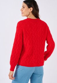 Una donna con capelli scuri indossa un maglione rosso a treccia e jeans azzurri chiari, orientata leggermente a sinistra con le mani appoggiate vicino ai fianchi.