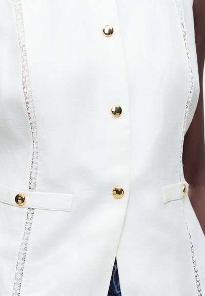 Gros plan sur un gilet blanc texturé avec des bordures en dentelle, des boutons ronds dorés et deux passants en tissu sur les côtés avant.