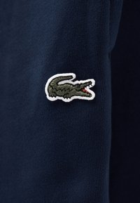 Πράσινο κεντητό λογότυπο κροκοδείλου με λευκό περίγραμμα σε σκούρο μπλε ύφασμα, τοποθετημένο στο μανίκι ενός ρούχου.