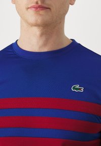 Homme portant un t-shirt bleu à col rond avec deux bandes rouges horizontales et un petit logo de crocodile vert sur la poitrine gauche.