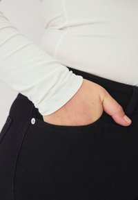 Schwarze Jeans mit einer Seitentasche und einem Knopfverschluss. Ein weißes Langarmshirt ist sichtbar, mit glatter, enganliegender Textur.