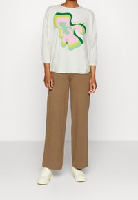 Ljust grå långärmad topp med en flerfärgad abstrakt design i rosa och grönt; kombinerad med lösa bruna stickade byxor och vita sneakers.