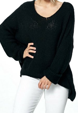 Mujer que lleva un suéter de punto negro suelto con mangas largas y pantalones blancos, con una mano en la cadera y la otra relajada hacia abajo.