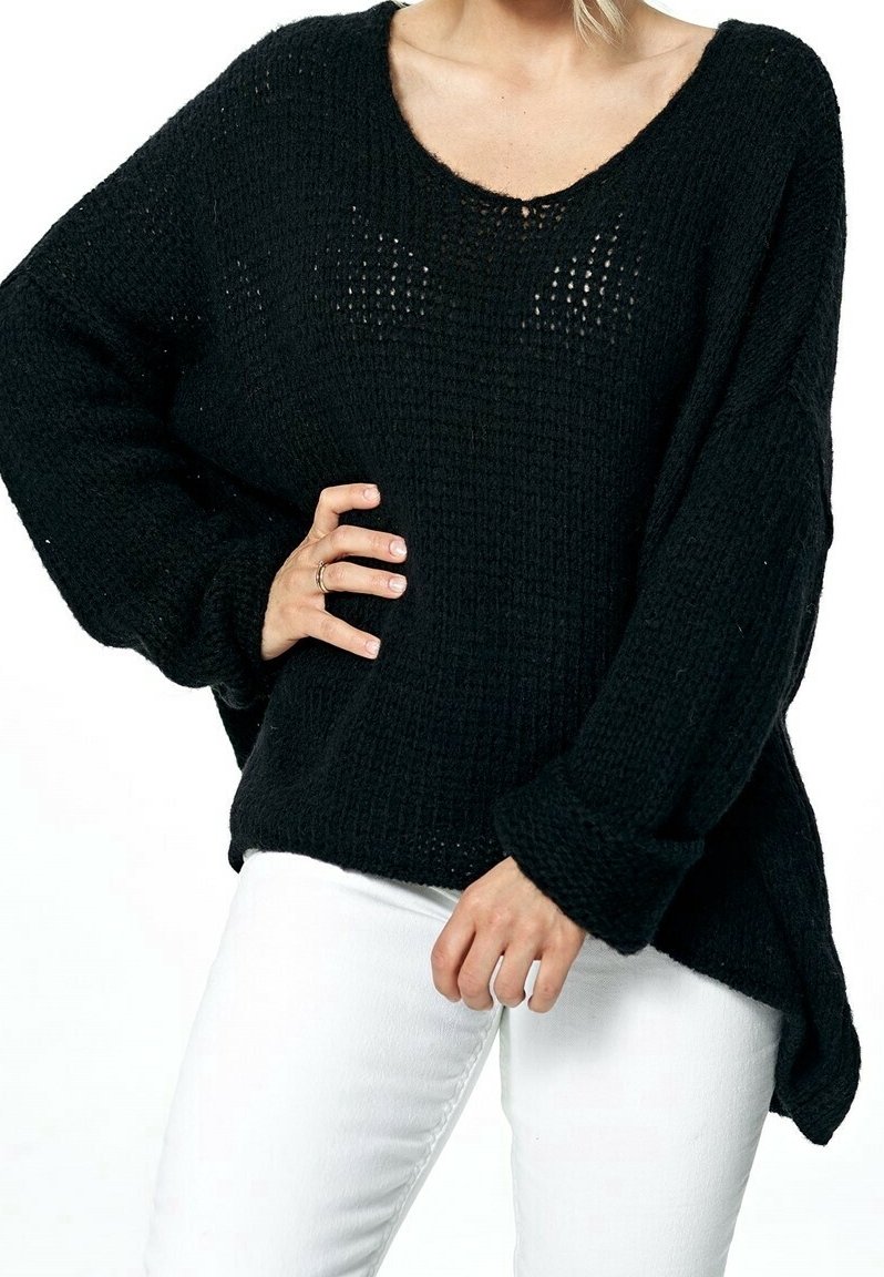 Femme portant un pull en maille noir ample à manches longues et un pantalon blanc, une main sur la hanche et l'autre détendue le long du corps.
