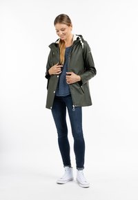 Olivenfarvet vandtæt jakke med hætte, med hvide snore og sidelommer, båret over en navy-stribet skjorte og blå jeans.