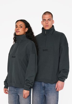 Sweatshirts gris foncé à col zippé quart, avec une coupe oversize, des poignets côtelés et le logo "Multiply Apparel" sur la poitrine. Portés avec un jean bleu.
