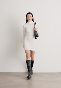 Rochie albă cu mâneci lungi și gât înalt. Botine negre până la genunchi și o mică poșetă neagră. Textură netedă cu detalii de design minime.