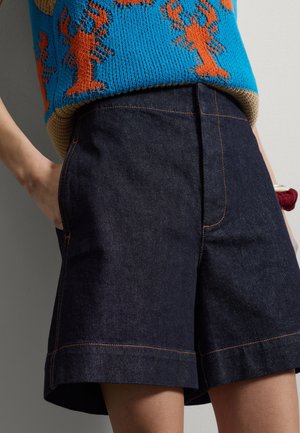 Pantalones cortos vaqueros oscuros de talle alto con anchos puños enrollados, costuras naranjas, combinados con un top de punto azul con un patrón de langosta naranja.