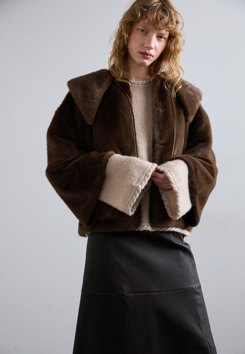 Giacca in pelliccia sintetica marrone con ampio colletto e maniche oversize, indossata sopra un maglione beige chiaro. La gonna in pelle nera completa l'outfit.