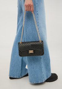 Borsa nera scintillante con motivo a zigzag, tracolla in catena dorata e dettaglio del clasp rettangolare. Indossata a mano contro jeans blu.