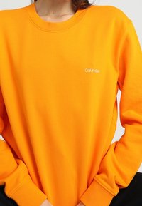 Jasnopomarańczowa bluza z zaokrąglonym dekoltem i długimi rękawami. Na piersi małe białe logo Calvin Klein. Miękki, teksturowany materiał.
