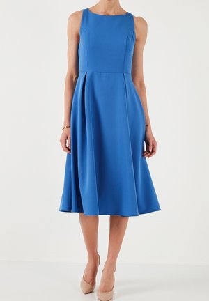 Mouwloze blauwe jurk tot op de knie met een aansluitend lijfje en een uitlopende rok, gedragen met naakte puntige hoge hakken en een gouden armband.