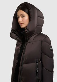 Tummanruskea toppatakki, jossa suuri hupullinen huppu, vetoketjullinen ja nepparikiinnitteinen, hihassa logo.