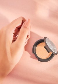 Fard à paupières bronze dans une palette ronde noire avec un couvercle transparent ; essai de la même teinte sur une main à la carnation claire.