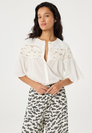 Frau trägt eine weiße Bluse mit Spitzeneinsätzen und weiten Ärmeln, kombiniert mit weißen Hosen mit schwarzem Fischmuster, steht vor schlichtem Hintergrund.