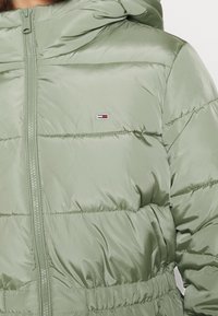 Prošívaná bunda světle zelené barvy s plným zipem, kapucí a elastickým pasem. Na levé straně hrudi je malý logotyp.