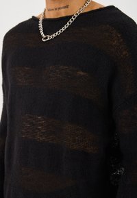 Schwarzer Strickpullover mit horizontalen durchsichtigen Streifen, entspannter Schnitt und weitem Ausschnitt. Dazu eine silberne Kettenkette.