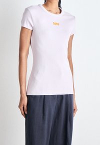 Ljusrosa figurnära t-shirt med korta ärmar, ribbad textur och liten gul "BOSS"-logga på framsidan. Buren med marinblå vida byxor.