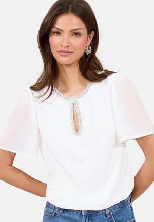 White blouse with short, puffed sleeves, featuring a round neckline and a keyhole cutout adorned with clear gemstones. Worn with blue jeans.