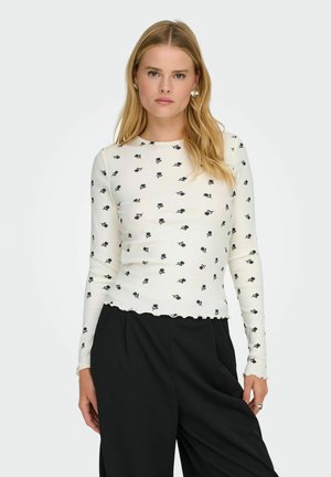 Witte geribde lange mouw top met een klein zwart bloemenpatroon en golvende zoom. Gecombineerd met zwarte losse pantalon.