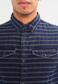 Camicia blu con bottoni realizzata in tessuto con finitura texturizzata, caratterizzata da righe orizzontali bianche, due tasche sul petto e bottoni a contrasto.