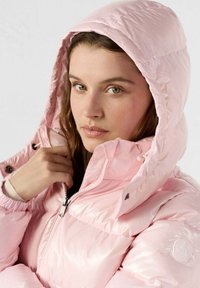 Giacca rosa gonfiata con finitura lucida, cappuccio staccabile, chiusura con zip e tasche laterali con logo rotondo sulla manica.
