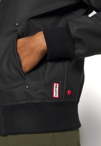 Ruka v kapse černé bundy Hunter s vroubkovaným lemem a spodním lemem, ukazující červené a bílé logo Hunter a detaily červeného patentního knoflíku.