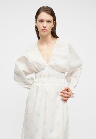 Γυναίκα με ίσια καστανά μαλλιά, που φοράει χρυσούς κρίκους στα αυτιά και λευκό φόρεμα με μπλε ανθισμένα σχέδια και φαρδιές μακριές μανίκες.