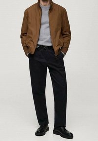 Homme portant une veste marron zippée, un pull gris, une ceinture noire et un pantalon noir, avec des chaussures habillées noires, debout les mains dans les poches.