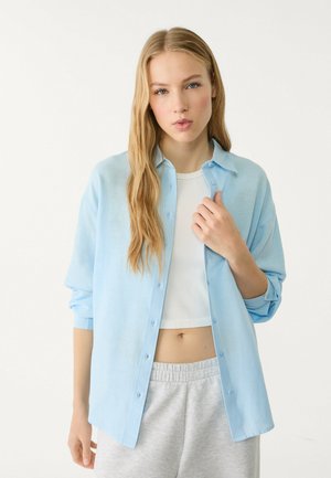 Light blue button-down shirt in soft fabric with long sleeves, worn over a white crop top and paired with gray sweatpants.