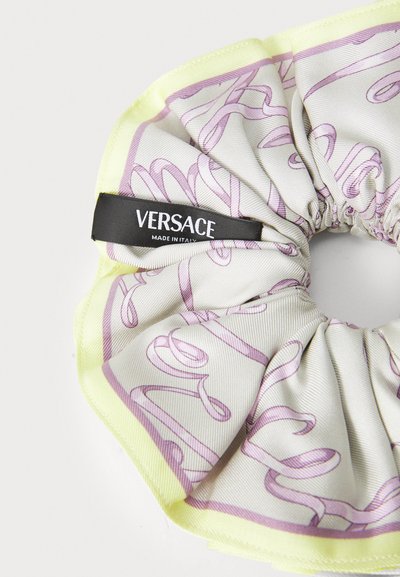 Svila scrunchie z bledo rumenim robom in vijoličastim abstraktnim vzorcem, ki ima črno oznako Versace z napisom "Made in Italy".