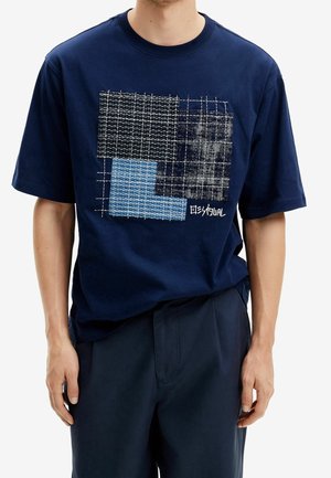 Mand iført mørkeblå T-shirt med gitter- og patchworkmønster i sorte, grå og blå firkanter på brystet.