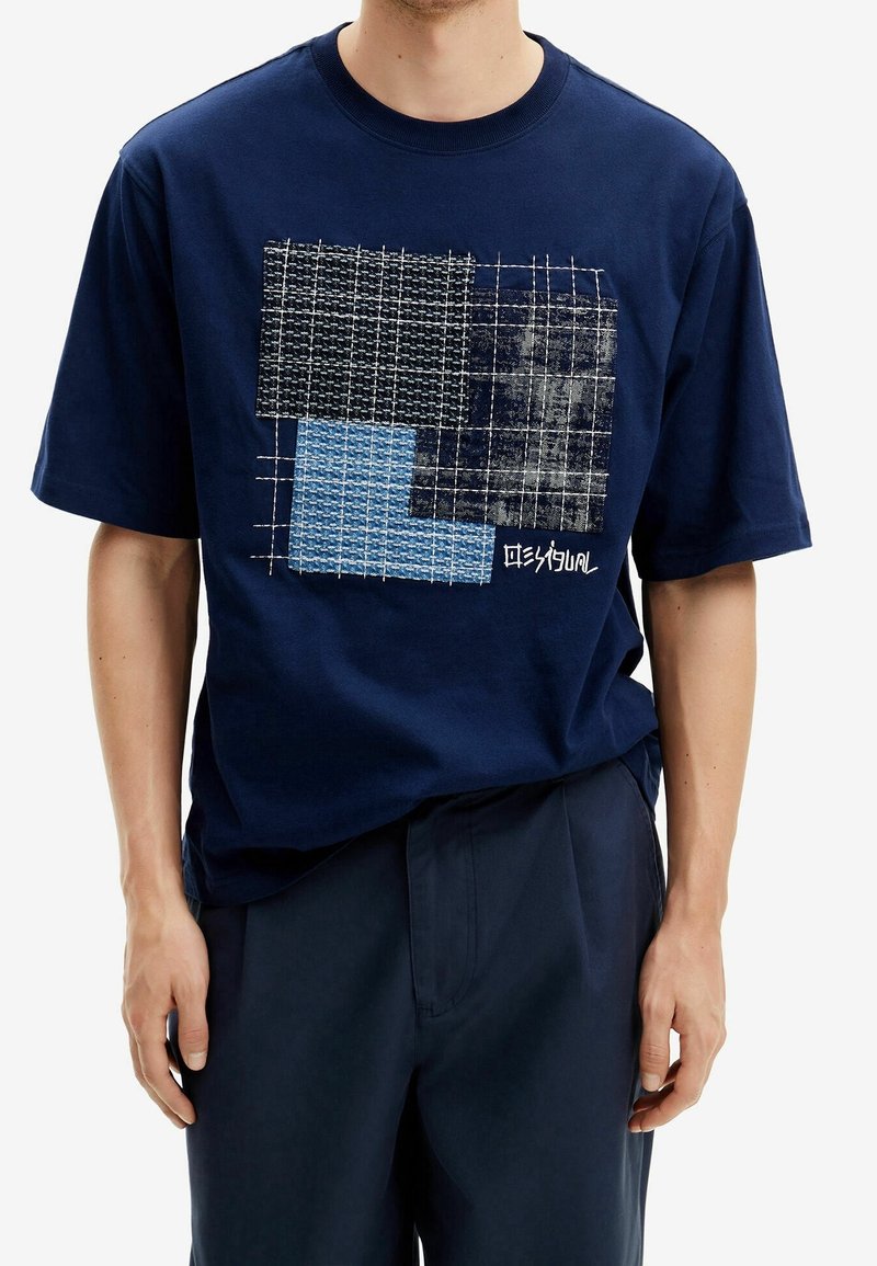 Homme portant un T-shirt bleu marine avec un motif en quadrillage et patchwork en carrés noirs, gris et bleus sur la poitrine.