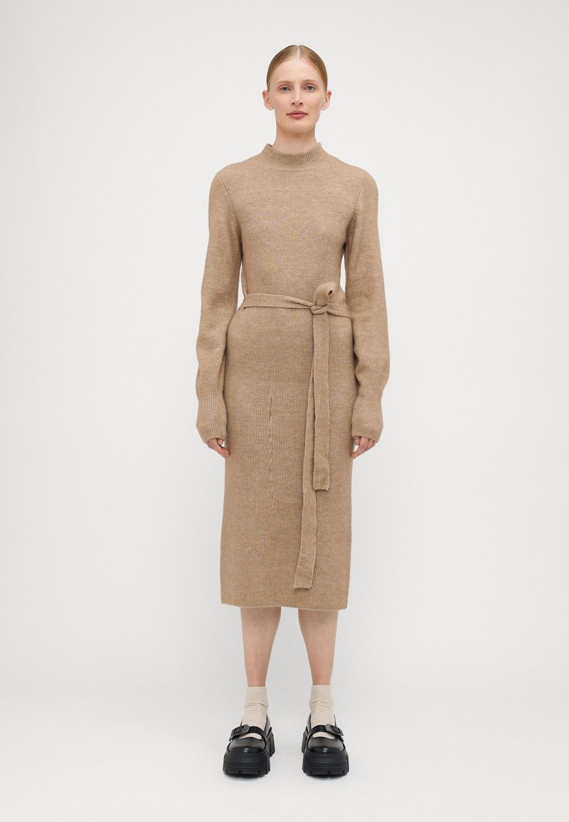 Abito in maglia marrone chiaro con collo alto, maniche lunghe e cintura legata. Presenta una texture a coste e un design al ginocchio. Scarpe con plateau nere.