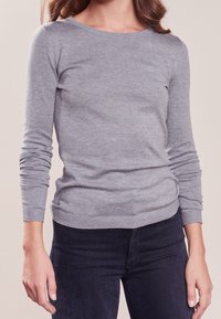 Kobieta ubrana w dopasowany, jasnoszary sweter z długim rękawem i ciemnoniebieskie dżinsy, stojąca na jednolitym beżowym tle.