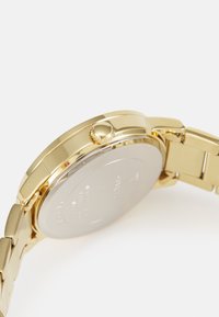 Orologio in metallo color oro con cassa circolare, corona testurizzata e bracciale a maglie. Il retro è di colore argento con testo inciso.