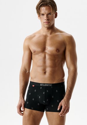 Muskularny mężczyzna o mokrych blond włosach prezentuje czarne bokserki Atlantic z drobnym powtarzającym się wzorem logo na białym tle.