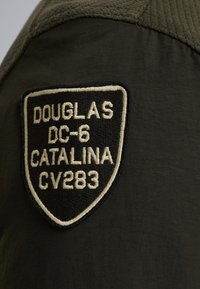 Tissu vert olive avec un écusson brodé noir en forme de bouclier portant le texte "DOUGLAS DC-6 CATALINA CV283" en lettres crème.