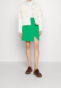Giacca di jeans bianca con frange, camicia bianca con colletto, gonna avvolgente verde e zoccoli marroni con suola simile al legno. Un outfit informale con texture sovrapposte.