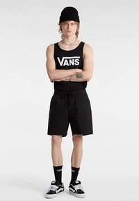 Schwarzes Vans Tanktop mit weißem Logo, schwarze Shorts mit Kordelzug, schwarze Mütze und Vans Schuhe. Accessoires umfassen schwarze Socken mit weißem Logo.