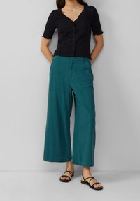 Black short-sleeve button-up top with a scalloped hem, paired with teal wide-leg pants and black strappy sandals.