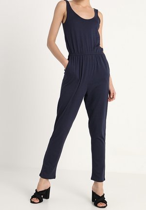 Kvinde iført en ærmeløs navy buksedragt med elastik i taljen og lommer, kombineret med sorte sandaletter med hæl, stående foran en ensfarvet baggrund.