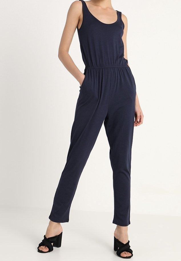 Femme portant une combinaison sans manches bleu marine avec taille élastique et poches, associée à des sandales à talons noires, debout devant un fond uni.