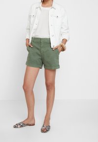 Olivově zelené bavlněné šortky se bočními kapsami, doplněné bílou džínovou bundou a vzorovanými sandály s křížovým designem.