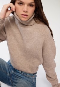 Jasno beżowy sweter z golfem, wykonany z miękkiego, teksturowanego materiału, z prążkowanym kołnierzem i luźnym krojem, zestawiony z niebieskimi dżinsami.