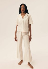 Kremowy zestaw koszuli z krótkim rękawem i spodni dzwonowatych, z jasnymi pionowymi paskami. Miękka tkanina o luźnym kroju i designie koszuli na guziki.