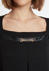 Schwarzes Oberteil mit quadratischem Ausschnitt, ausgestattet mit einem Satinband und einem dekorativen goldfarbenen Hardware-Detail in der Mitte. Einfaches und elegantes Design.