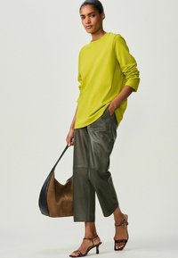 Limoengroene langoe sleeve top, ruime pasvorm, gecombineerd met donkergroene leren cropped broeken. Model houdt een twee-tonen schoudertas vast en draagt gespenhakken.