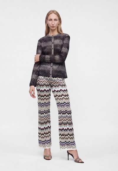 Femme debout, les bras croisés, portant un haut noir à manches longues à motifs et un pantalon multicolore à motif zigzag avec des sandales à talons hauts.